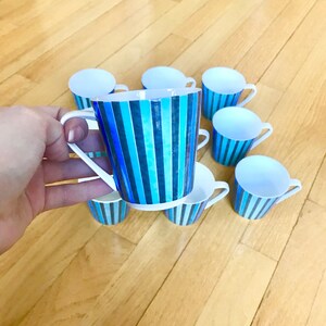 May include: A set of eight small white teacups with a vertical striped pattern in shades of blue. The cups have white handles and are arranged on a light wooden surface. One cup is held in a hand.