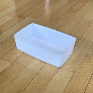 May include: A white plastic rectangular storage bin with a smooth surface. The bin is empty and sitting on a wooden floor.