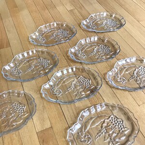 May include: Set of eight clear glass serving plates with a raised floral design. The plates are oval shaped with a scalloped edge.