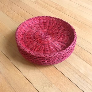 May include: A round, woven wicker basket with a pink stain. The basket is made of natural materials and has a rustic, handmade look.