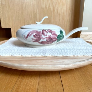 May include: A white ceramic serving dish with a lid and handle, decorated with pink and purple roses and green leaves. The dish is placed on a white, textured doily and a wooden tray.