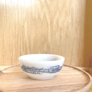 May include: A small, white ceramic bowl with a detailed gray illustration of a train and landscape. The bowl sits on a round, wooden serving tray. The background is a light-colored wooden surface.