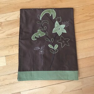 May include: A brown table runner with a green floral embroidery design. The runner has a green border.