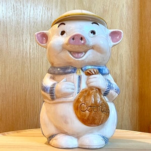 May include: A white ceramic cookie jar shaped like a pig wearing a blue and white striped shirt and a baseball cap. The pig is holding a brown bag that says "Cookies".