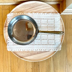 May include: A silver frying pan with a brass handle sits on a white crocheted placemat on a wooden tray.
