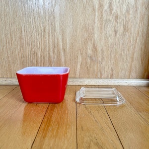 May include: A red glass storage container with a clear glass lid. The container is square shaped and has a white rim. The lid is slightly domed.