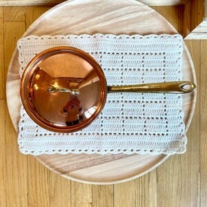May include: A copper saucepan with a lid and a brass handle rests on a cream-colored crocheted potholder. The potholder is placed on a round wooden tray. The scene is captured from an overhead perspective, showcasing the kitchenware.