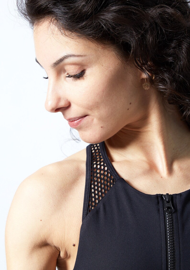 May include: A woman wearing a black athletic top with a mesh panel on the shoulder and a full-length zipper. The woman has dark curly hair and is wearing gold earrings. The background is white.