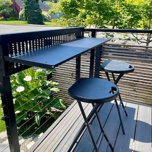 Op de afbeelding: Zwarte balkonset voor buiten met een klaptafel en twee bijpassende krukken. De tafel is op een reling gemonteerd en de krukken hebben een ronde zitting met een handvat. De set is gemaakt van metaal en kunststof.