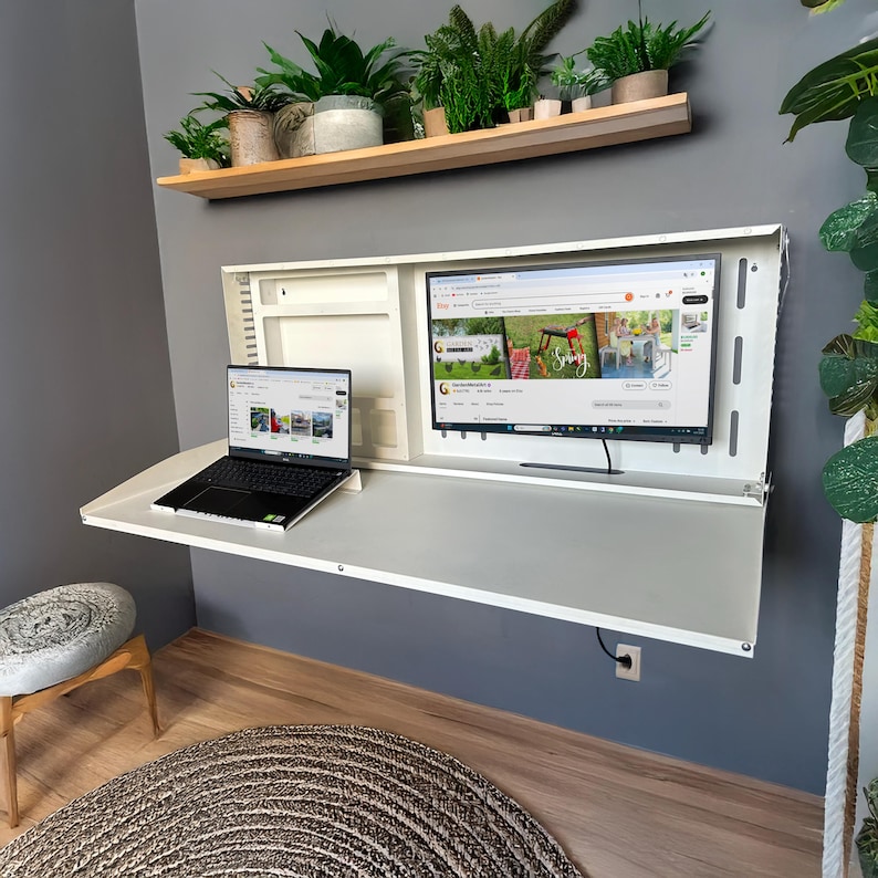 May include: A white, wall-mounted desk with a built-in monitor and a laptop. The desk is open, revealing a workspace. A wooden shelf above holds potted plants. The monitor displays a website. The room has a gray wall and wooden floor.
