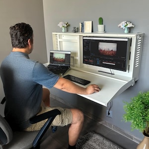 May include: A white wall-mounted desk with a built-in shelf, holding a laptop, a large monitor displaying video editing software, and a keyboard. The desk is open, and a person is seated in front of it.