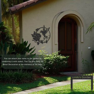 May include: Black metal hummingbird and flower garden decoration with a circular design. The decoration is mounted on a beige house exterior with a brown wooden door. The text "Linda's Garden" is visible on a black metal sign in the foreground.