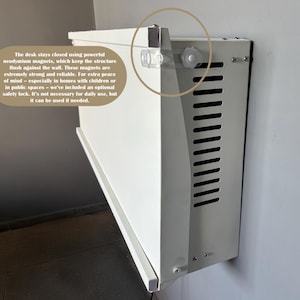 May include: A white, wall-mounted desk with a rectangular design. The desk features a series of ventilation slots and a safety lock mechanism. The text on the image states the desk uses neodymium magnets to stay closed.