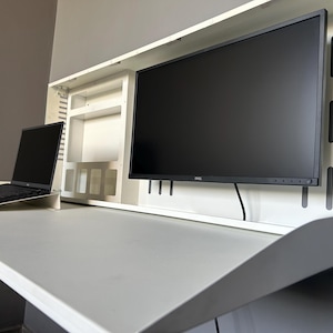 May include: A white, wall-mounted desk with a built-in monitor and a laptop. The desk has a fold-down design, with a large monitor and a laptop on the desk surface. The monitor is black, and the laptop is open.