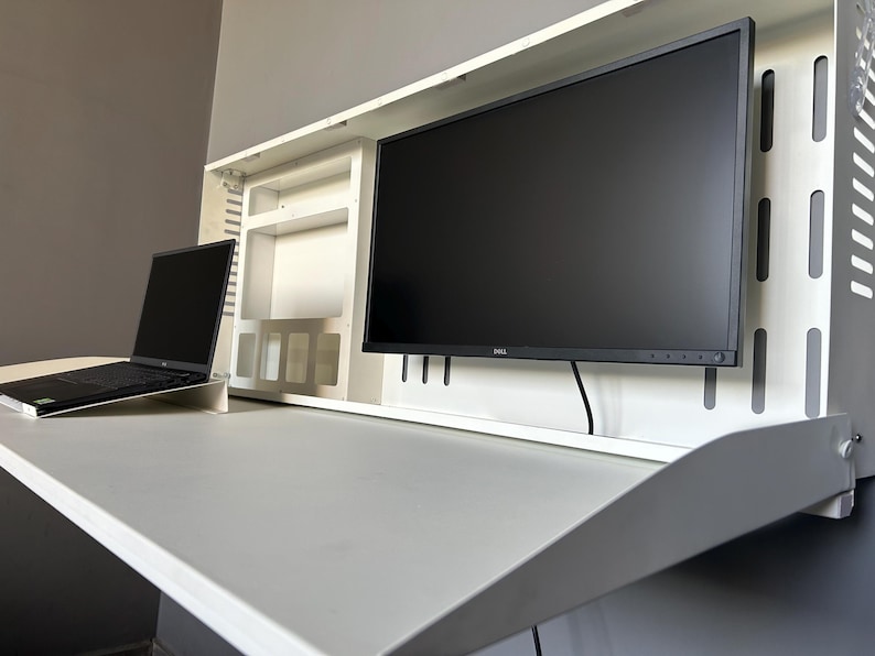 May include: A white, wall-mounted desk with a built-in monitor and a laptop. The desk has a fold-down design, with a large monitor and a laptop on the desk surface. The monitor is black, and the laptop is open.