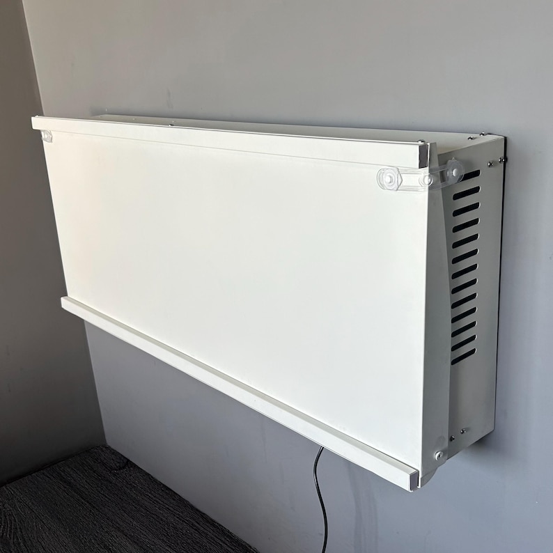 May include: A white rectangular wall-mounted cabinet with a slatted side panel. The cabinet is mounted on a gray wall and has a clear plastic corner piece. A black cord hangs down from the bottom.