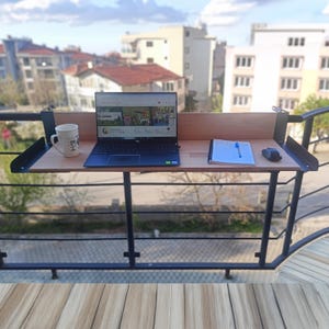 Peut inclure: Un bureau de balcon en bois avec un cadre en métal noir, contenant un ordinateur portable, une tasse, un bloc-notes et une souris. L'ordinateur affiche un site web. Le bureau est fixé à une balustrade en métal noir.
