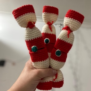 Puede incluir: Tres adornos de cracker de Navidad de ganchillo en rojo y blanco con detalles verdes. Los crackers están atados con hilo blanco.