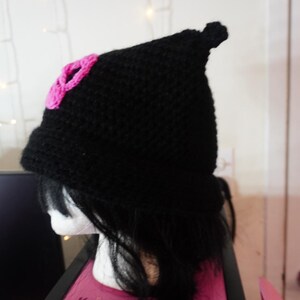 K&ouml;nnte beinhalten: Eine schwarze H&auml;kelm&uuml;tze mit einer rosa Blume an der Seite. Die M&uuml;tze hat eine spitze Spitze und wird auf einer Puppe getragen.