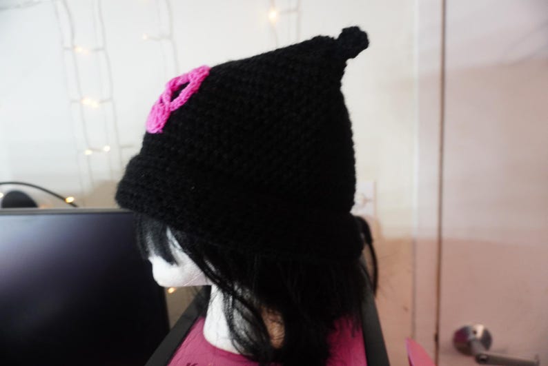 K&ouml;nnte beinhalten: Eine schwarze H&auml;kelm&uuml;tze mit einer rosa Blume an der Seite. Die M&uuml;tze hat eine spitze Spitze und wird auf einer Puppe getragen.