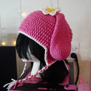K&ouml;nnte beinhalten: Rosa gestrickte M&uuml;tze mit wei&szlig;em Rand und einer wei&szlig;en Blume oben. Die M&uuml;tze hat lange Ohrenklappen, die unter dem Kinn zusammengebunden werden.