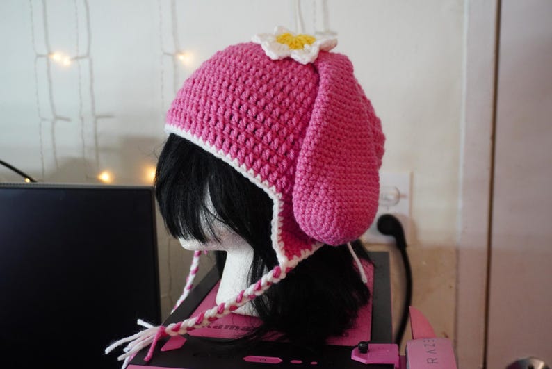 K&ouml;nnte beinhalten: Rosa gestrickte M&uuml;tze mit wei&szlig;em Rand und einer wei&szlig;en Blume oben. Die M&uuml;tze hat lange Ohrenklappen, die unter dem Kinn zusammengebunden werden.