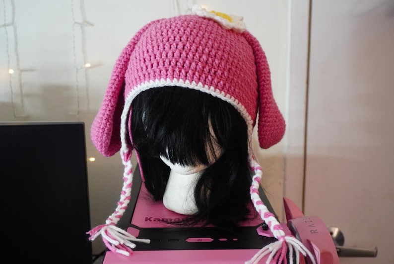 K&ouml;nnte beinhalten: Rosa gestrickte M&uuml;tze mit wei&szlig;em Besatz und Hasenohren. Die M&uuml;tze hat eine wei&szlig;e Blume oben drauf.
