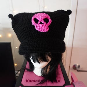 K&ouml;nnte beinhalten: Schwarze H&auml;kelm&uuml;tze mit einem rosafarbenen Totenkopf-Design. Die M&uuml;tze hat zwei kleine Ohrenklappen.