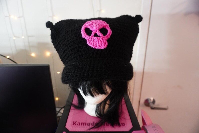 K&ouml;nnte beinhalten: Schwarze H&auml;kelm&uuml;tze mit einem rosafarbenen Totenkopf-Design. Die M&uuml;tze hat zwei kleine Ohrenklappen.