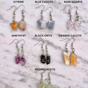 May include: A set of 10 pairs of earrings featuring different types of crystals, including citrine, blue calcite, rose quartz, amethyst, black onyx, orange calcite, and green calcite. Each pair of earrings features a crystal wrapped in silver wire and attached to a silver hook.