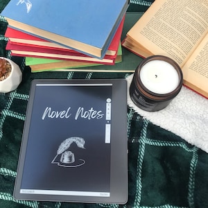 May include: A black e-reader displaying the words "Novel Notes" on the screen. The e-reader is on a green and white checked blanket with a stack of books and a candle.