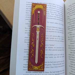 May include: A burgundy bookmark with a sword design. The bookmark features a detailed illustration of a sword with a gold hilt and blade, set against a burgundy background with gold accents. The bookmark is rectangular and is placed on top of an open book.