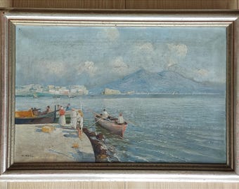 Op de afbeelding: Olieverfschilderij in een zilveren lijst dat een kustlandschap uitbeeldt. Het kunstwerk toont boten, mensen, gebouwen en een berg op de achtergrond. Het kleurenpalet omvat blauw, groen en bruin.