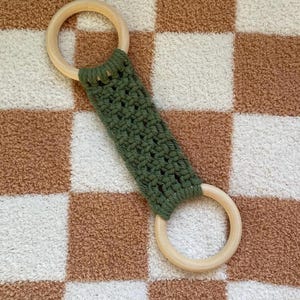 May include: A green crocheted teething toy with two wooden rings. The toy is on a brown and white checkered surface.