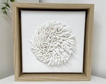 May include: A white framed canvas with a white 3D textured design of a circle with raised white lines. The design resembles a flower or coral.