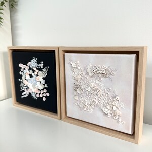 May include: Two framed square canvases with abstract floral designs. The canvas on the left has a black background with white, pink, and blue floral shapes. The canvas on the right has a white background with white floral shapes.