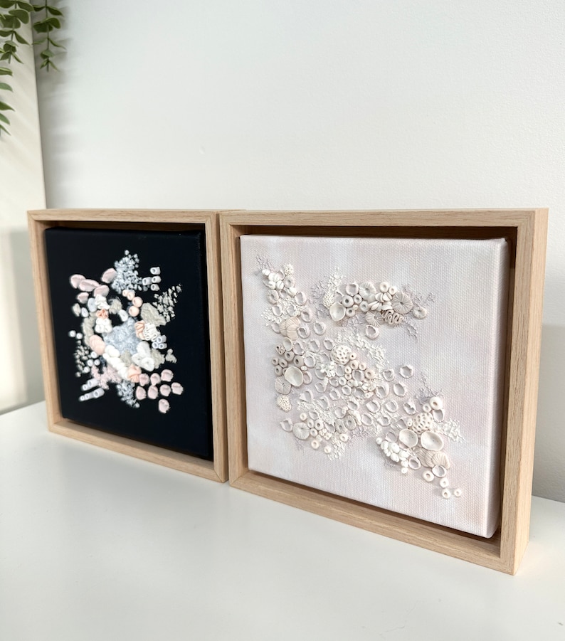 May include: Two framed square canvases with abstract floral designs. The canvas on the left has a black background with white, pink, and blue floral shapes. The canvas on the right has a white background with white floral shapes.