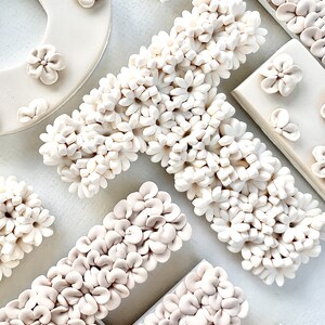 以下が含まれることがあります： 白いセラミック製の文字が、小さな白い花の形をした装飾で覆われています。文字は白い背景にジグザグに配置されています。