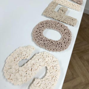 以下が含まれることがあります： ベージュと白のフェルトの花でできた「SOFA」という単語を綴る5つの文字のセット。文字は白い表面に一列に並んでいます。
