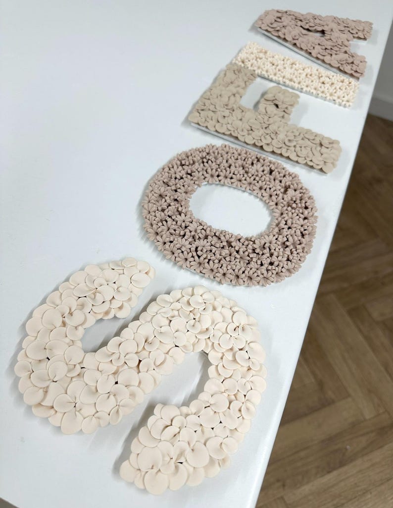 以下が含まれることがあります： ベージュと白のフェルトの花でできた「SOFA」という単語を綴る5つの文字のセット。文字は白い表面に一列に並んでいます。