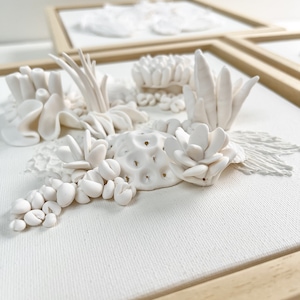 May include: Close-up of a white, textured artwork depicting a coral reef scene. The artwork is composed of various sculpted elements, including coral formations and plant-like structures, mounted on a white canvas within a light wooden frame. Several similar artworks are visible.