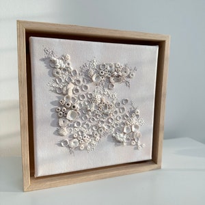 May include: A framed canvas with a white background and a 3D design of white shells and coral. The design is arranged in a cluster and appears to be made of clay or ceramic.