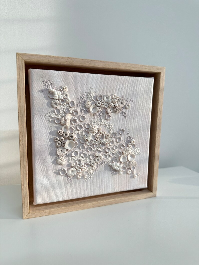 May include: A framed canvas with a white background and a 3D design of white shells and coral. The design is arranged in a cluster and appears to be made of clay or ceramic.