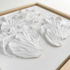 May include: White ceramic flower petals arranged in a wave pattern on a white canvas with a wooden frame. The petals are layered and have a delicate texture.