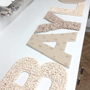 以下が含まれることがあります： 花柄のデザインで「BABY」と綴られたベージュと白の文字のセット。文字は質感のある素材でできており、白い表面に配置されています。