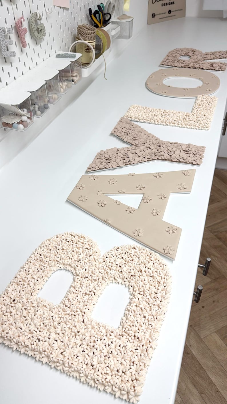 以下が含まれることがあります： 花柄のデザインで「BABY」と綴られたベージュと白の文字のセット。文字は質感のある素材でできており、白い表面に配置されています。