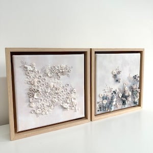May include: Two framed canvas paintings with a textured, 3D effect. The left painting features white and cream textured shapes, while the right painting features blue and gray textured shapes.