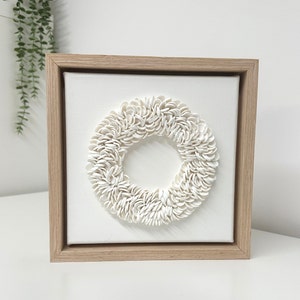 May include: A white ceramic wreath with a circular shape, framed in a light wood frame. The wreath is made of many small, overlapping pieces of ceramic, creating a textured and organic look.