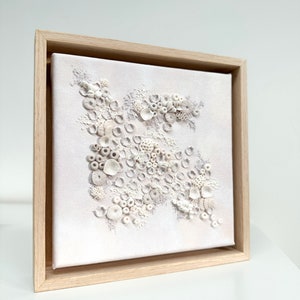 May include: A white framed canvas with a textured, raised design of white circles and shapes, resembling coral or seashells.