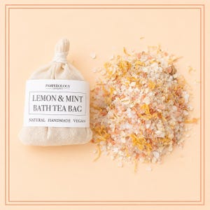May include: A beige fabric bath tea bag with a white label reading PAMPEROLOGY LONDON LEMON & MINT BATH TEA BAG NATURAL HANDMADE VEGAN, positioned next to a loose pile of pink Himalayan salt, dried yellow flower petals, and white oat flakes.